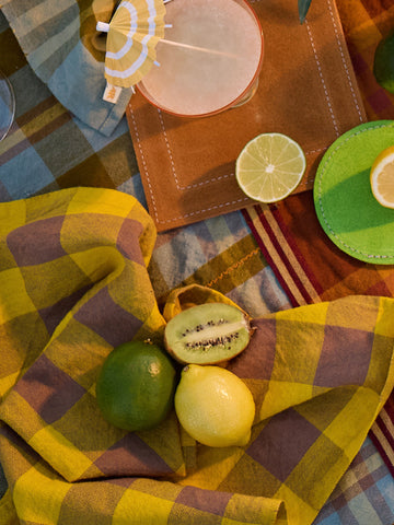 Fruits on a checkered tablecloth with colorful coasters and a drink.
