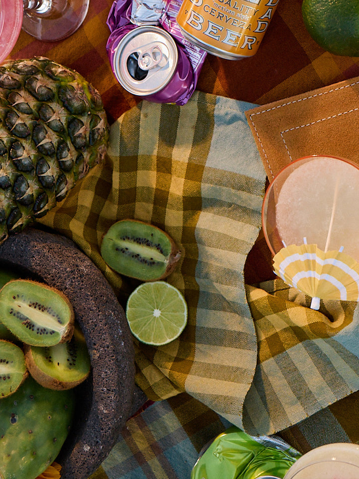 Table setting with pineapple, kiwi, lime, and beer on a checkered tablecloth.