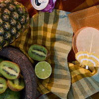 Table setting with pineapple, kiwi, lime, and beer on a checkered tablecloth.