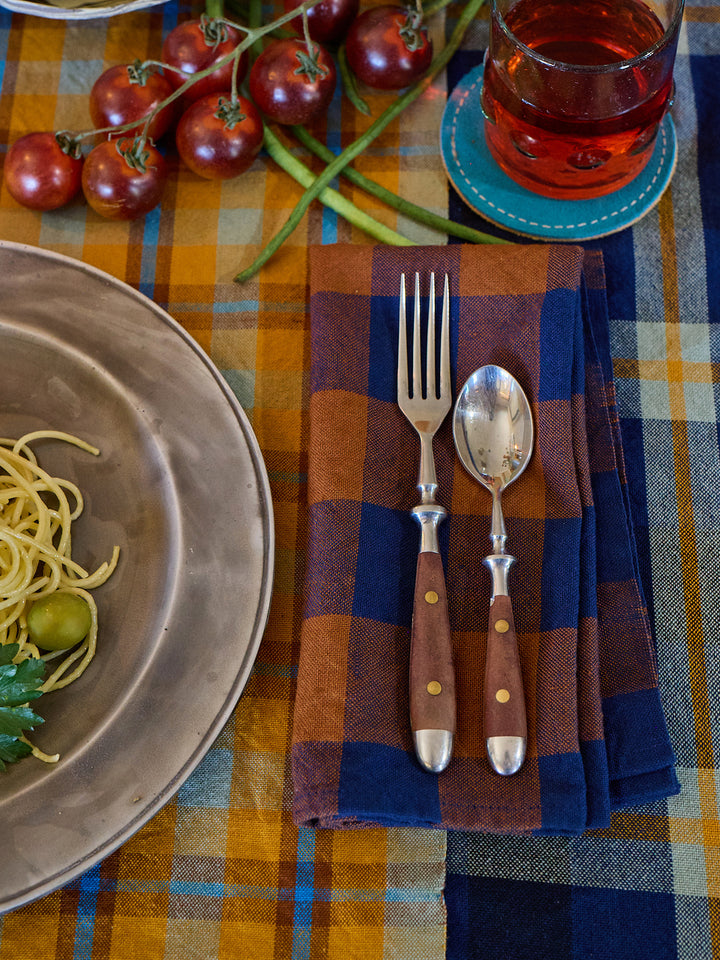 Dinner setting with a bowl of pasta, fork, spoon, and plaid napkin on a checkered tablecloth.