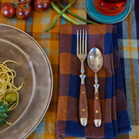 Dinner setting with a bowl of pasta, fork, spoon, and plaid napkin on a checkered tablecloth.