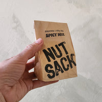 Hand holding a small brown paper bag labeled 'Spicy Mix Nut Sacks' against a light background
