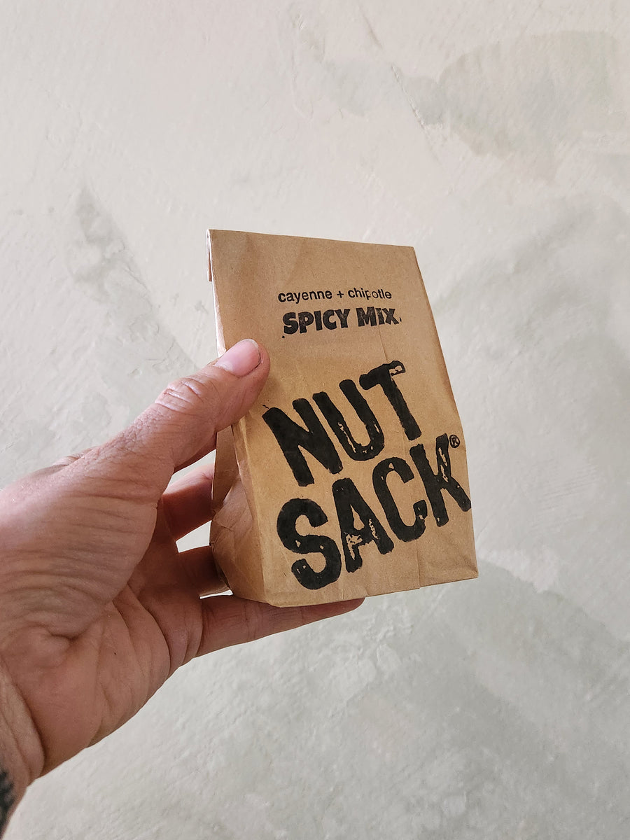 Hand holding a small brown paper bag labeled 'Spicy Mix Nut Sacks' against a light background