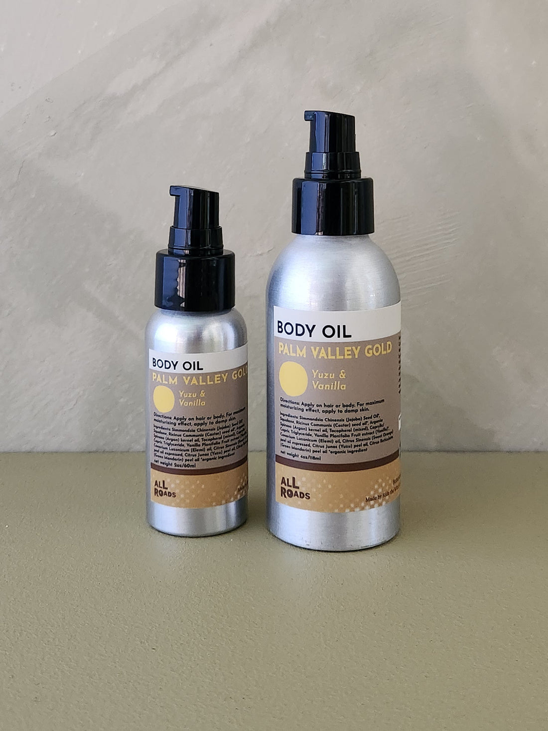 Two silver spray bottles of Palm Valley Gold body oil, one in a smaller pump bottle and one in a larger pump bottle, with black caps and beige labels, set against a neutral textured background.