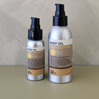 Two silver spray bottles of Palm Valley Gold body oil, one in a smaller pump bottle and one in a larger pump bottle, with black caps and beige labels, set against a neutral textured background.