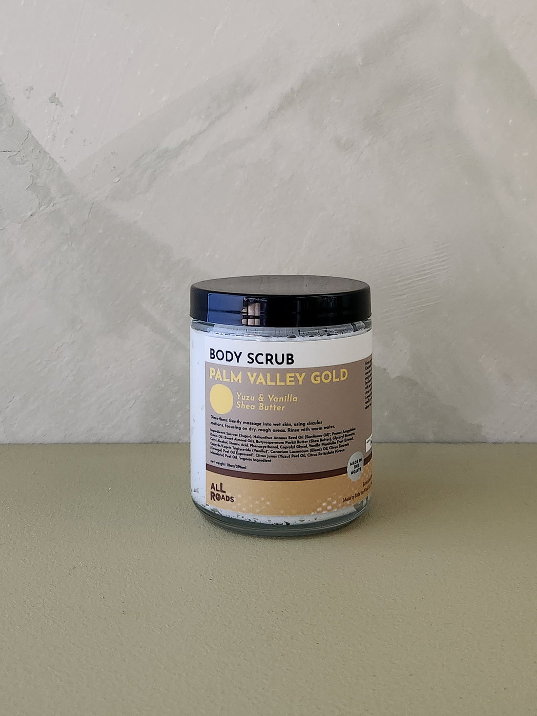 A jar of Palm Valley Gold body scrub by All Natural, featuring a black lid and a white and beige label, set against a neutral textured gray background.