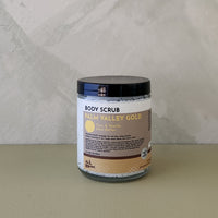 A jar of Palm Valley Gold body scrub by All Natural, featuring a black lid and a white and beige label, set against a neutral textured gray background.