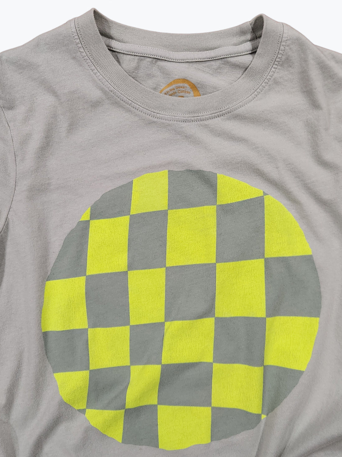 A close-up of a light sage green crewneck t-shirt featuring a large circular graphic on the front with a yellow and gray checkerboard pattern.
