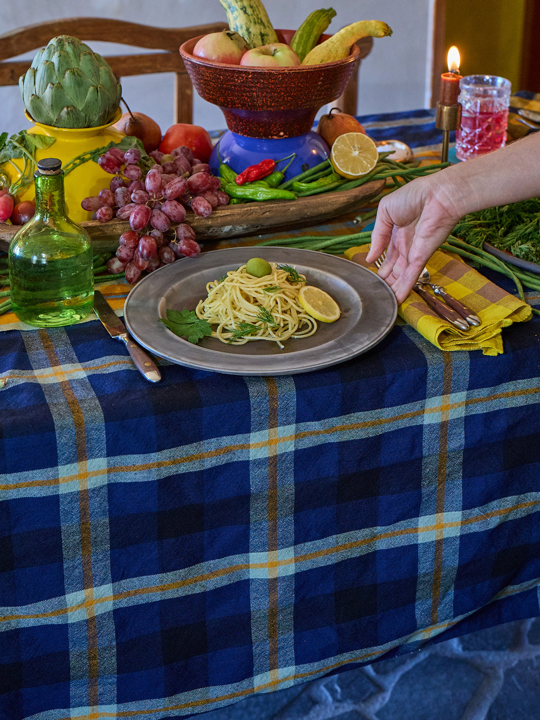 Table setting with pasta dish, fruit, and a candle on a blue plaid tablecloth.