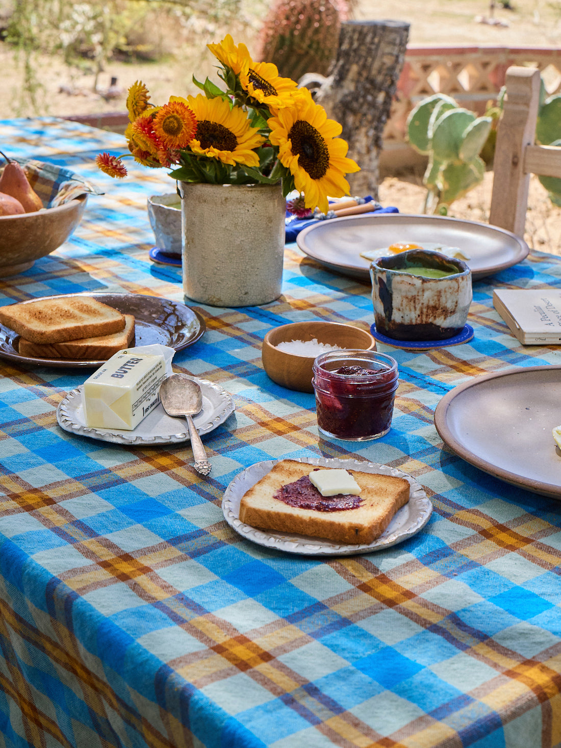Outdoor breakfast table with plaid tablecloth, sunflowers, and food items.