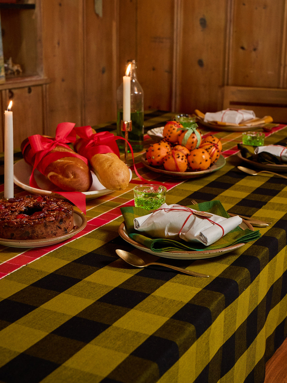 A rustic holiday table setting featuring a prominent yellow and black buffalo plaid tablecloth. The table is decorated with plates of fruit, bread rolls tied with red ribbons, lit taper candles, and wrapped gifts, set against a warm wooden background.