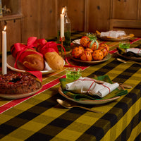 A rustic holiday table setting featuring a prominent yellow and black buffalo plaid tablecloth. The table is decorated with plates of fruit, bread rolls tied with red ribbons, lit taper candles, and wrapped gifts, set against a warm wooden background.
