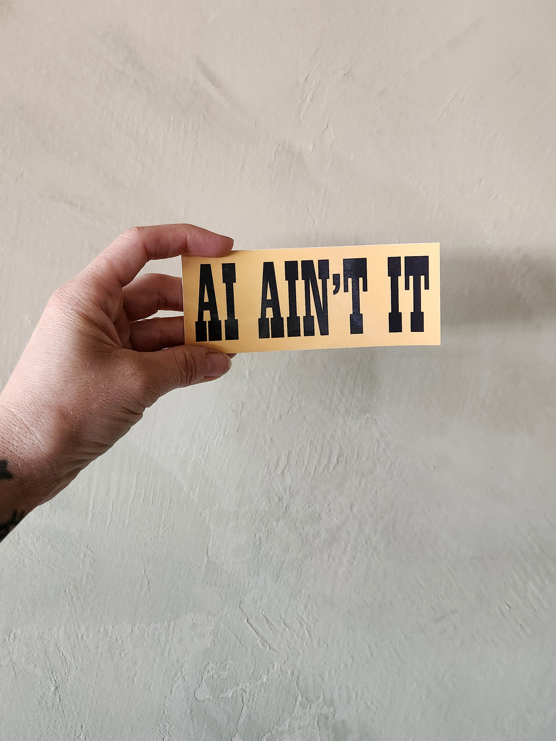 A hand holds a rectangular beige sticker or card with the phrase &