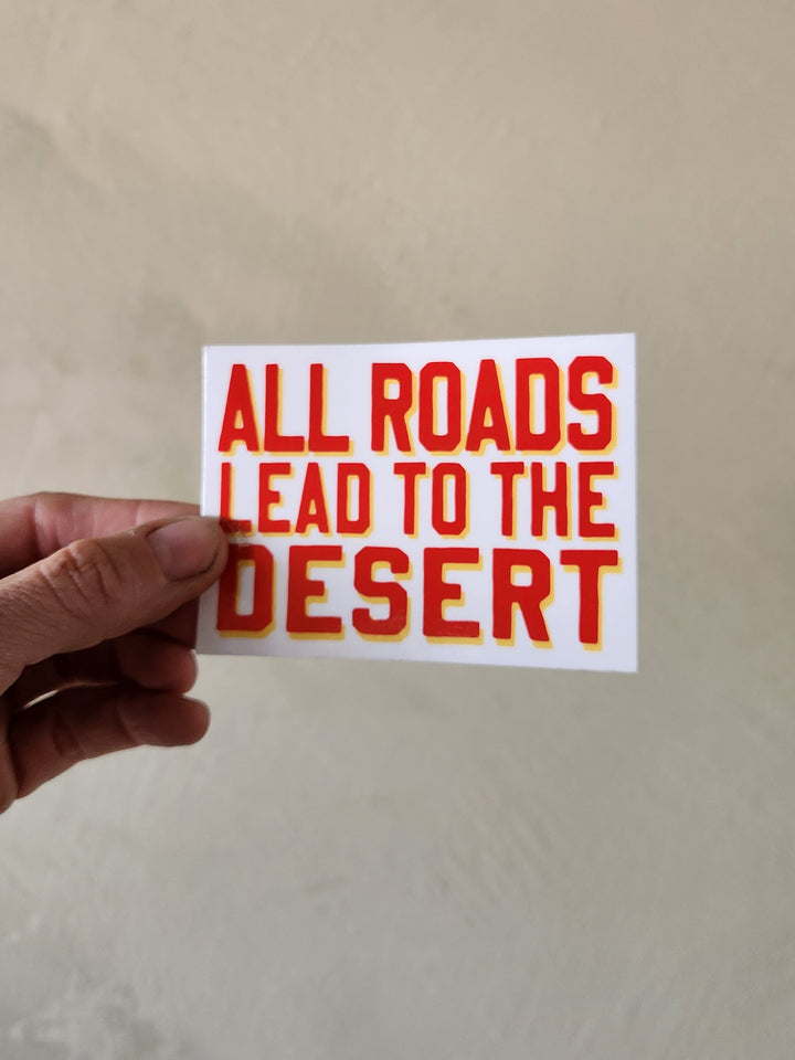 Hand holding a sticker with 'All roads lead to the desert' text on a plain background