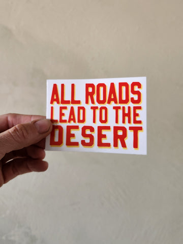 Hand holding a sticker with 'All roads lead to the desert' text on a plain background