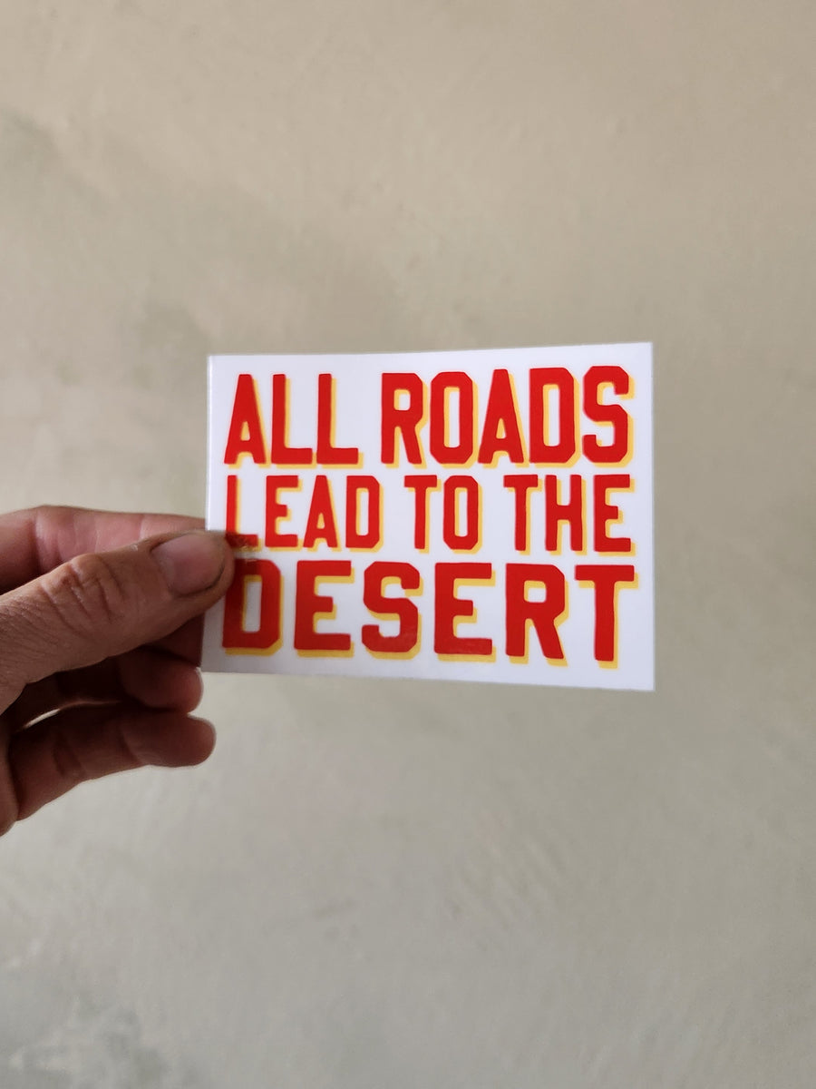 Hand holding a sticker with 'All roads lead to the desert' text on a plain background