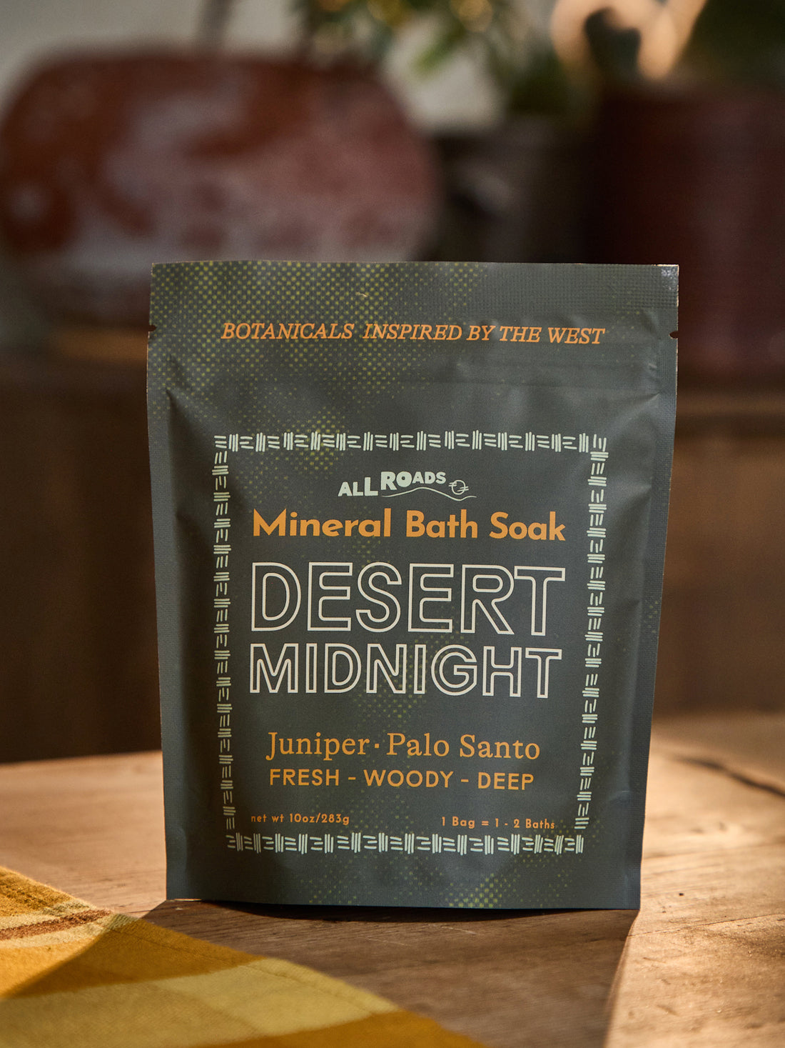 A stand-up pouch of All Roads Mineral Bath Soak in the scent Desert Midnight, placed on a wooden surface with a warm, blurred background.