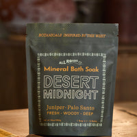 A stand-up pouch of All Roads Mineral Bath Soak in the scent Desert Midnight, placed on a wooden surface with a warm, blurred background.