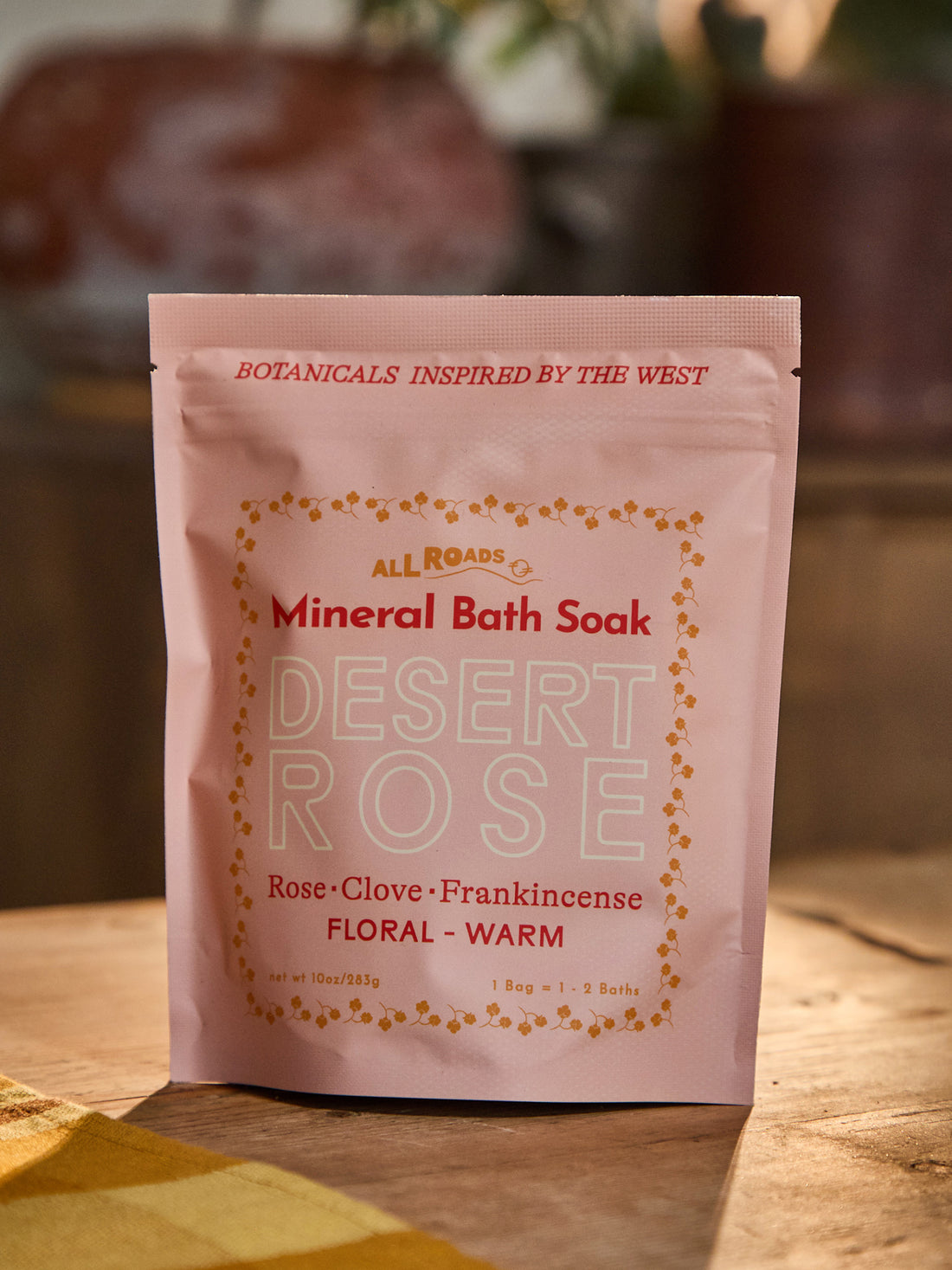 A pink stand-up pouch of All Roads Botanicals Mineral Bath Soak in the Desert Rose scent, placed on a wooden surface with a warm, blurred background.