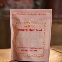 A pink stand-up pouch of All Roads Botanicals Mineral Bath Soak in the Desert Rose scent, placed on a wooden surface with a warm, blurred background.