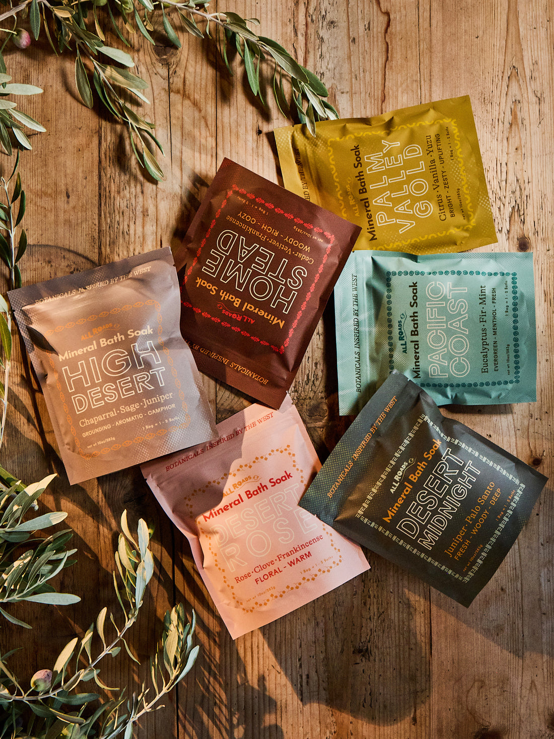 Six colorful pouches of Mineral Bath Soak in various flavors and colors are arranged in a flat-lay on a rustic wooden table with olive branches.