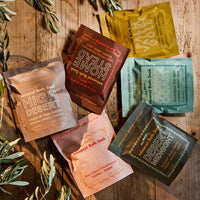 Six colorful pouches of Mineral Bath Soak in various flavors and colors are arranged in a flat-lay on a rustic wooden table with olive branches.