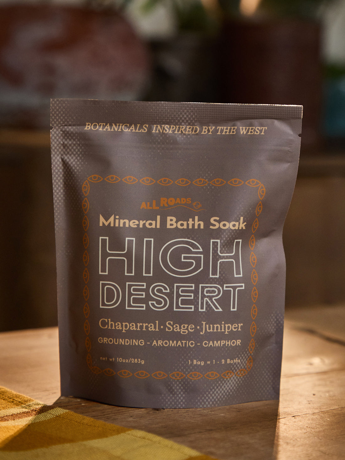 A stand-up pouch of All Roads Mineral Bath Soak in the High Desert scent, placed on a wooden surface with a warm, blurred background.