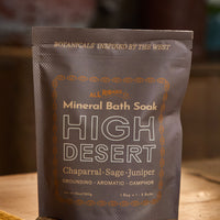A stand-up pouch of All Roads Mineral Bath Soak in the High Desert scent, placed on a wooden surface with a warm, blurred background.