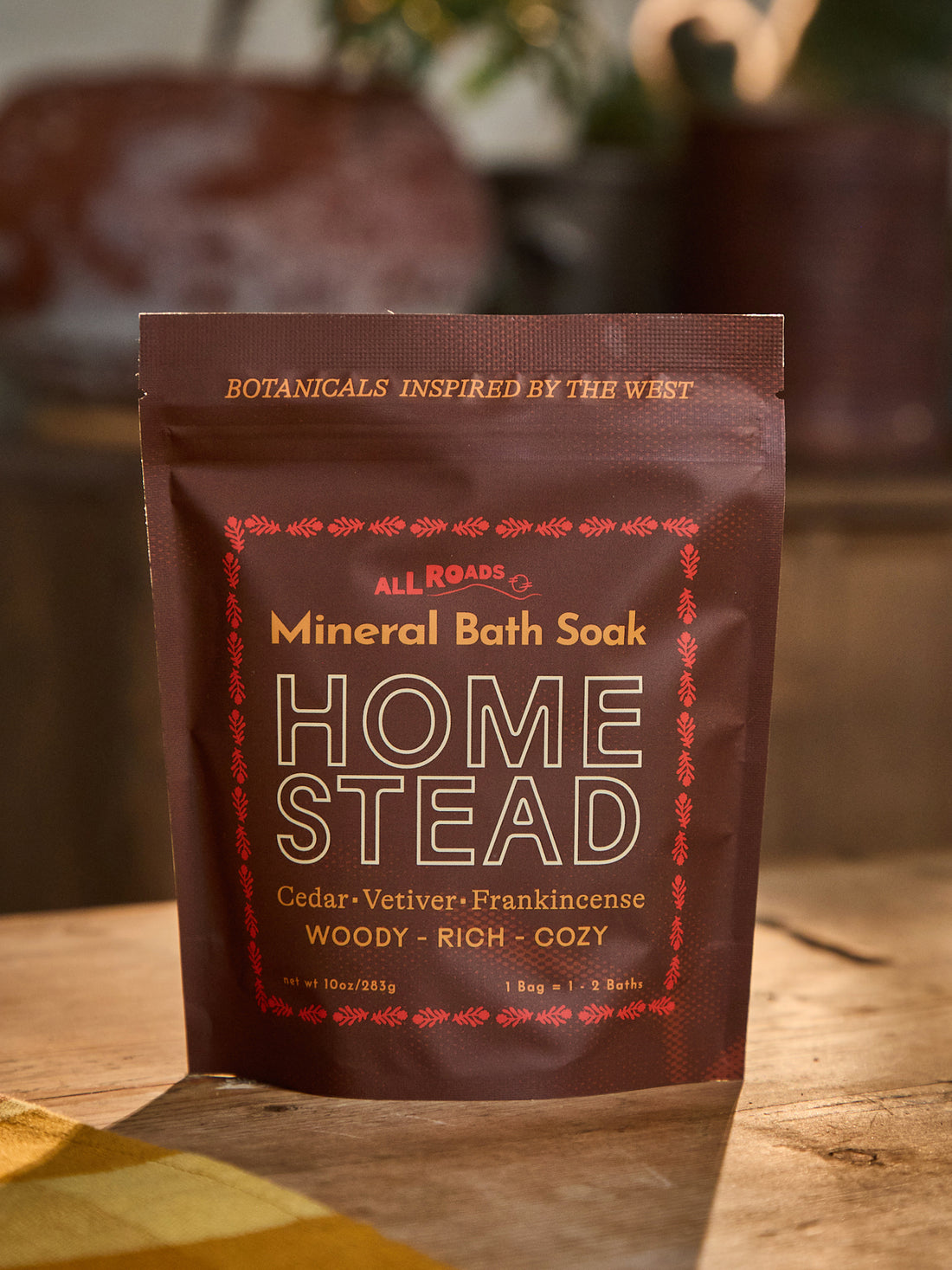 A stand-up pouch of All Roads Homestead Mineral Bath Soak, featuring a dark brown color with gold and red text and decorative borders, set on a wooden surface with a warm, blurred background.
