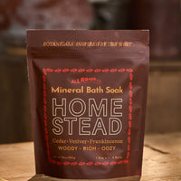 A stand-up pouch of All Roads Homestead Mineral Bath Soak, featuring a dark brown color with gold and red text and decorative borders, set on a wooden surface with a warm, blurred background.