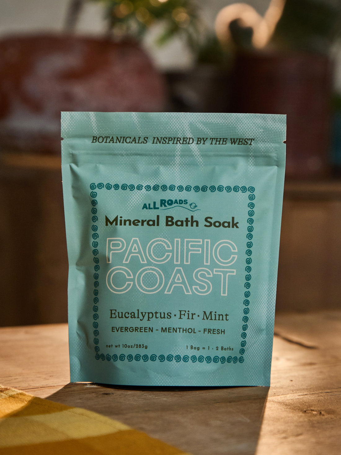 A stand-up pouch of All Roads Mineral Bath Soak, flavor Pacific Coast (Eucalyptus, Fir, Mint), standing on a wooden surface with a warm, blurred background.