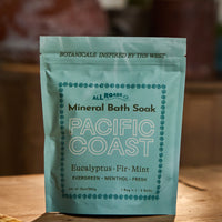 A stand-up pouch of All Roads Mineral Bath Soak, flavor Pacific Coast (Eucalyptus, Fir, Mint), standing on a wooden surface with a warm, blurred background.