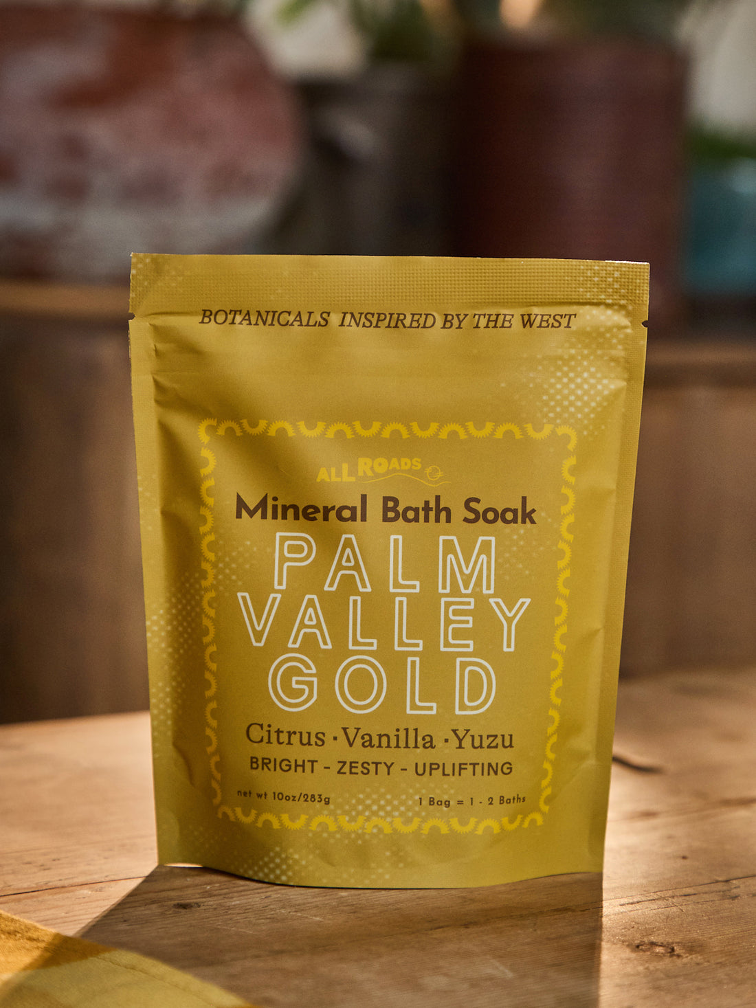 A stand-up pouch of Palm Valley Gold Mineral Bath Soak by All Roads, featuring a golden-yellow design with black and white text, set on a wooden surface with a blurred rustic background.