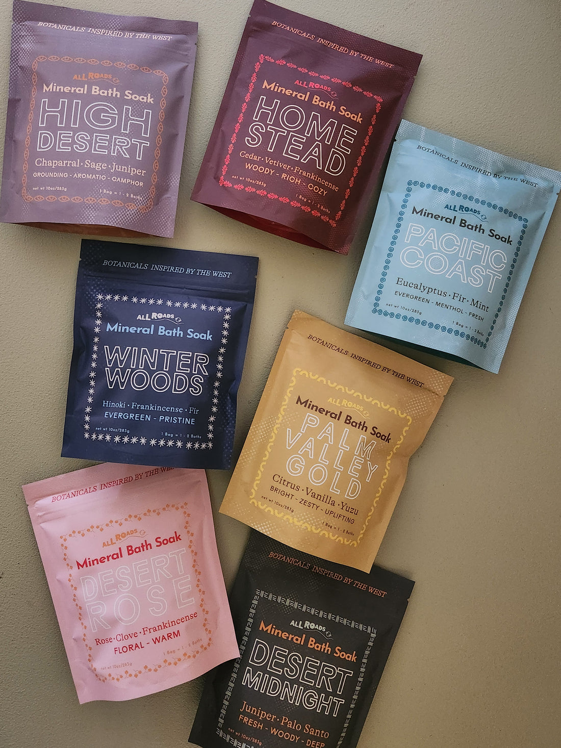 Seven pouches of botanical-inspired mineral bath soaks in assorted scents, arranged in a flat-lay pattern on a neutral beige background. The pouches come in a variety of colors including purple, burgundy, light blue, navy, mustard yellow, pink, and black, each with its own unique illustrated label.