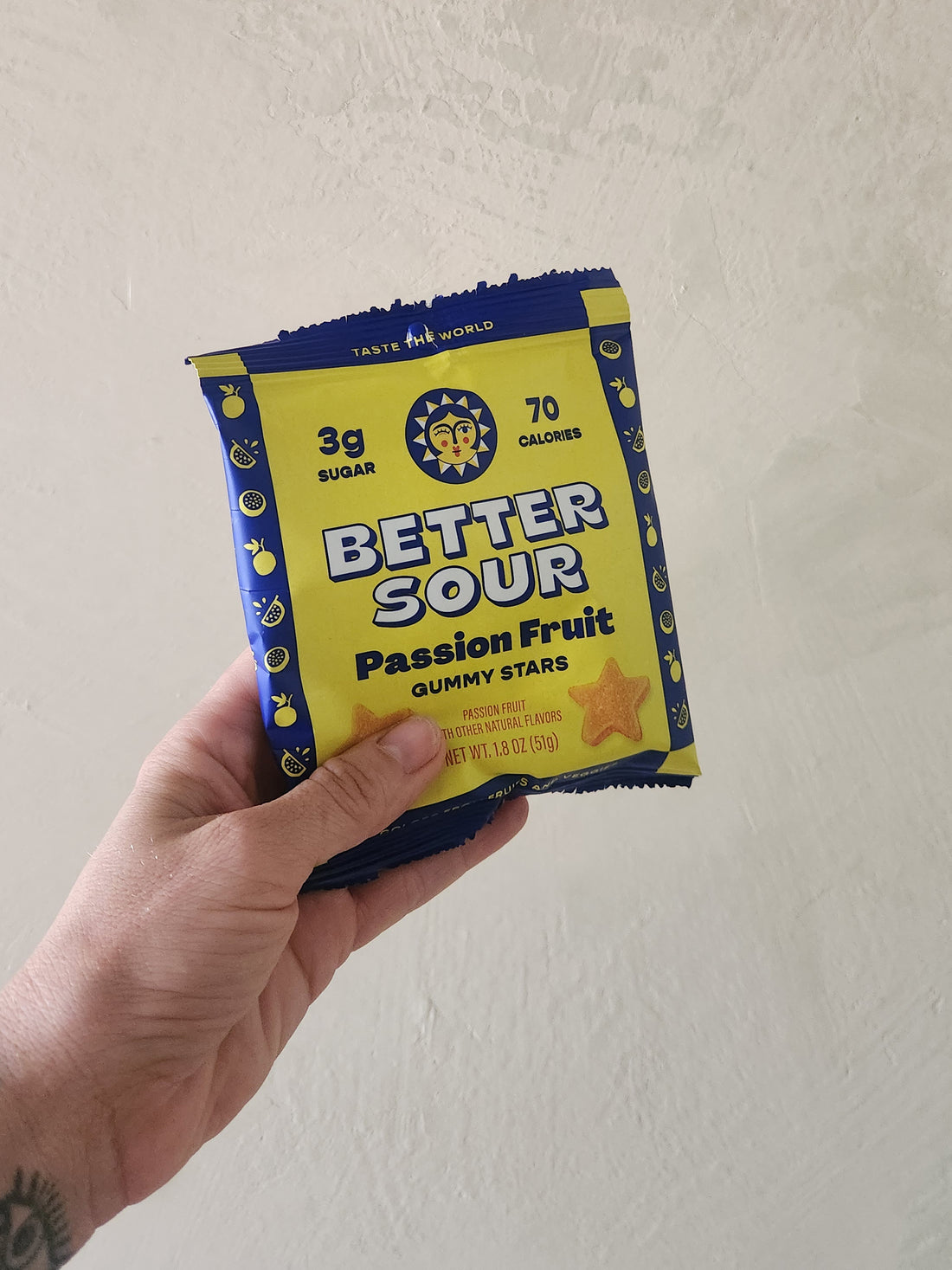 A hand holding a package of Better Sour Passion Fruit Gummy Stars against a plain beige wall.
