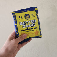 A hand holding a package of Better Sour Passion Fruit Gummy Stars against a plain beige wall.