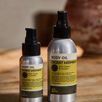 Two silver spray bottles of Body Oil by All Roads, one smaller and one larger, labeled 'DESERT MIDNIGHT' with yellow labels, set on a wooden surface with a blurred background.