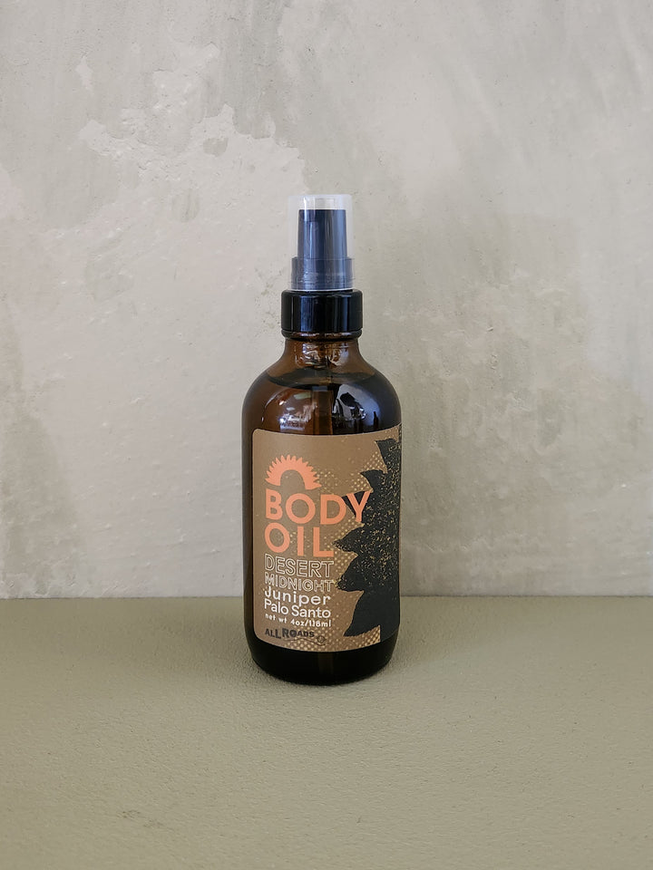 A bottle of Desert Midnight body oil with a spray nozzle, featuring a label with desert-inspired artwork.