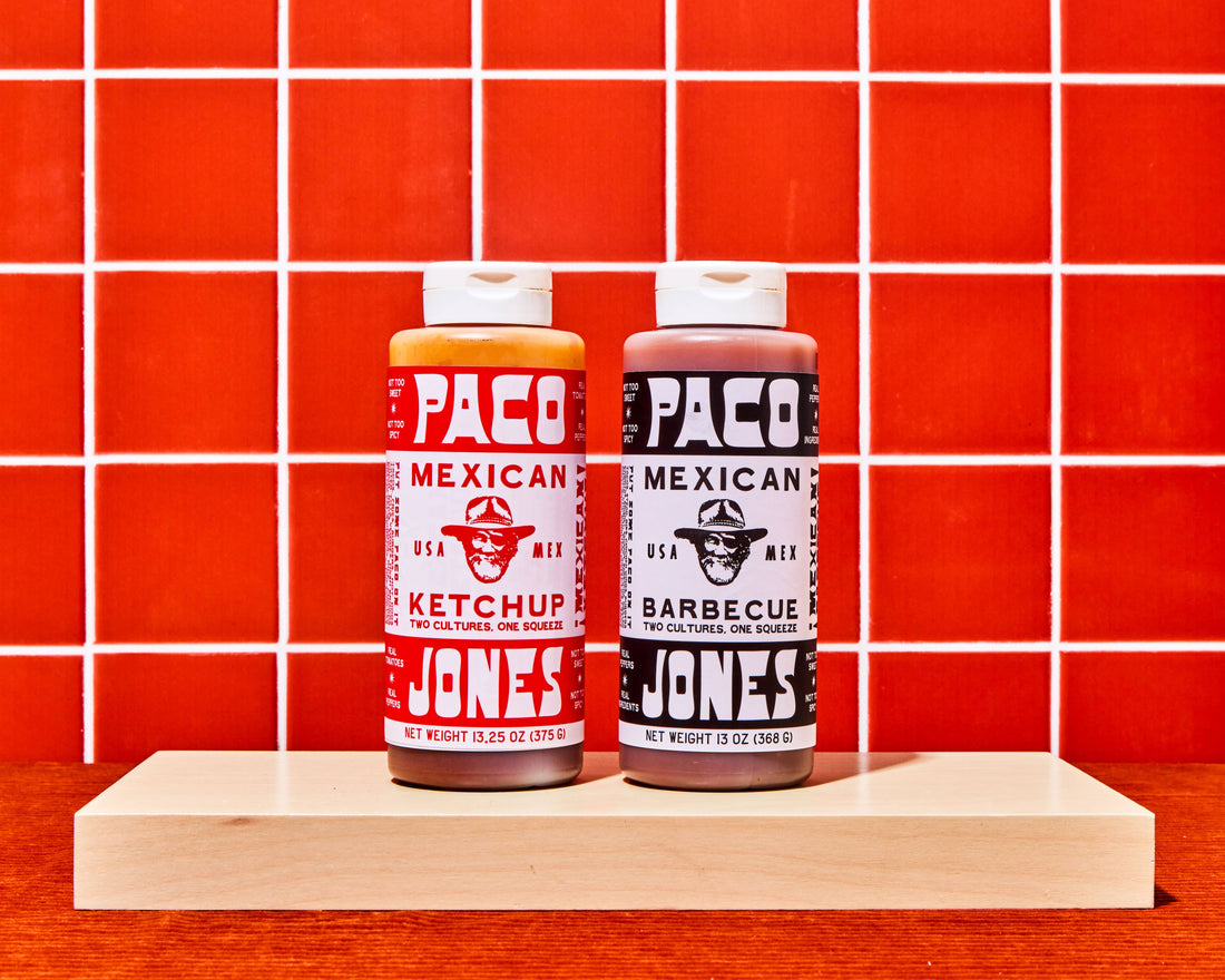Two bottles of taco seasoning from Paco Jones Mexican Ketchup and Barbecue Seasoning displayed side-by-side on a light wood block against a red tiled background.