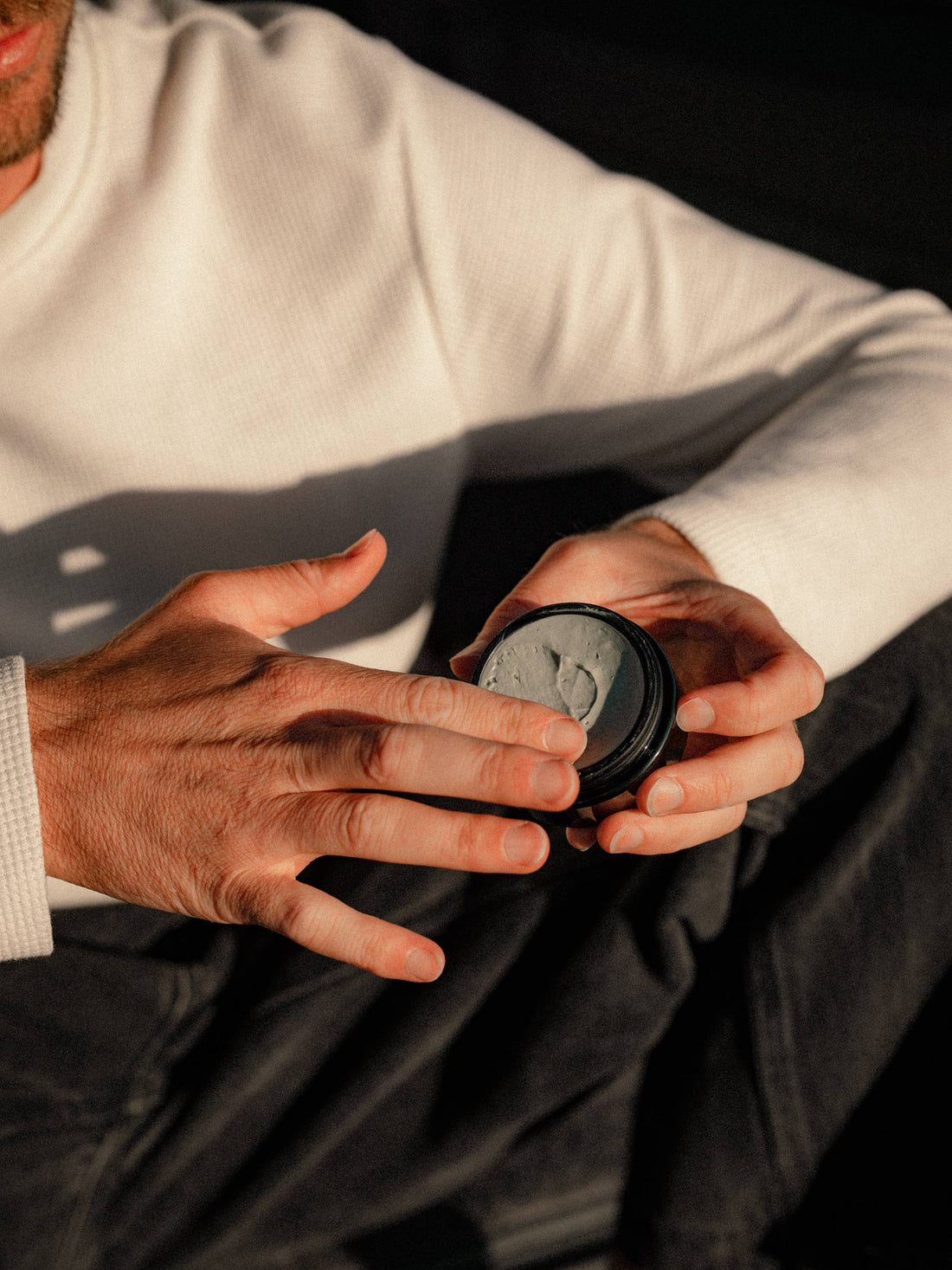 A close-up of a person holding an open round black jar containing a gray cream or balm, with their fingers touching the surface. The person is wearing a white ribbed sweater and dark pants, set against a dark background with dramatic warm lighting.