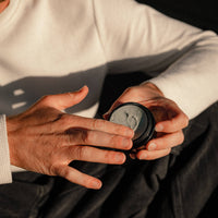 A close-up of a person holding an open round black jar containing a gray cream or balm, with their fingers touching the surface. The person is wearing a white ribbed sweater and dark pants, set against a dark background with dramatic warm lighting.