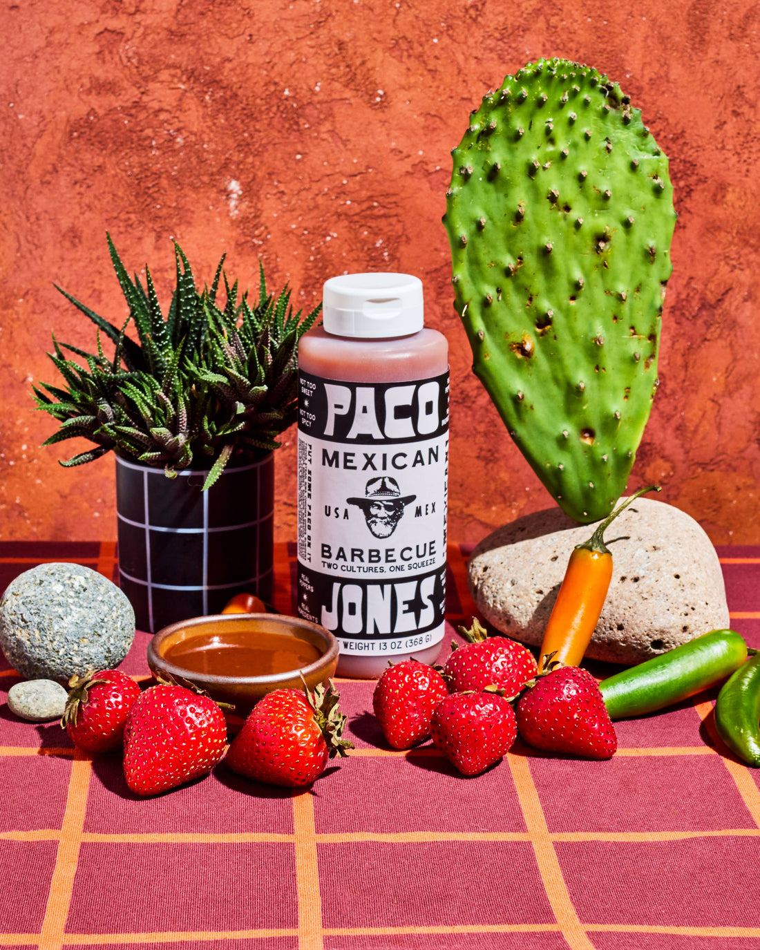 A bottle of Paco Mexican Barbecue sauce stands on a red plaid surface with strawberries, a small bowl of sauce, a cactus, a potted succulent, and other produce against a textured terracotta background.