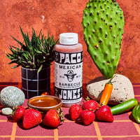 A bottle of Paco Mexican Barbecue sauce stands on a red plaid surface with strawberries, a small bowl of sauce, a cactus, a potted succulent, and other produce against a textured terracotta background.