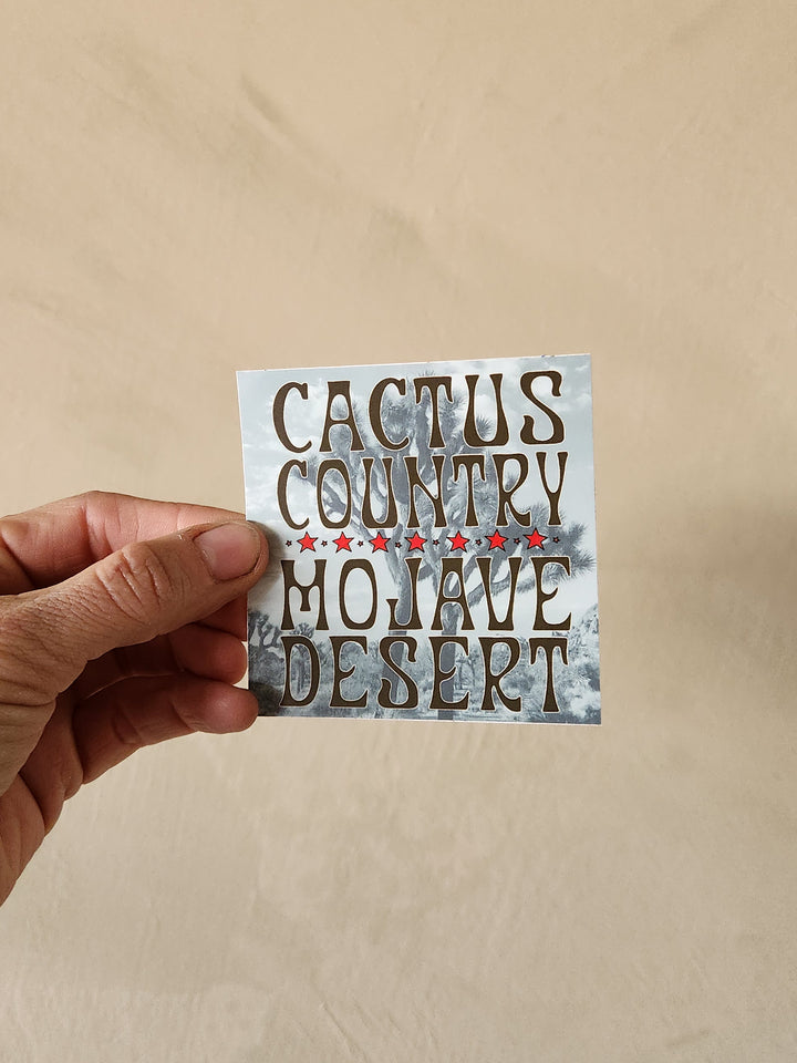 Hand holding a sticker with 'Cactus Country Mojave Desert' text against a beige background