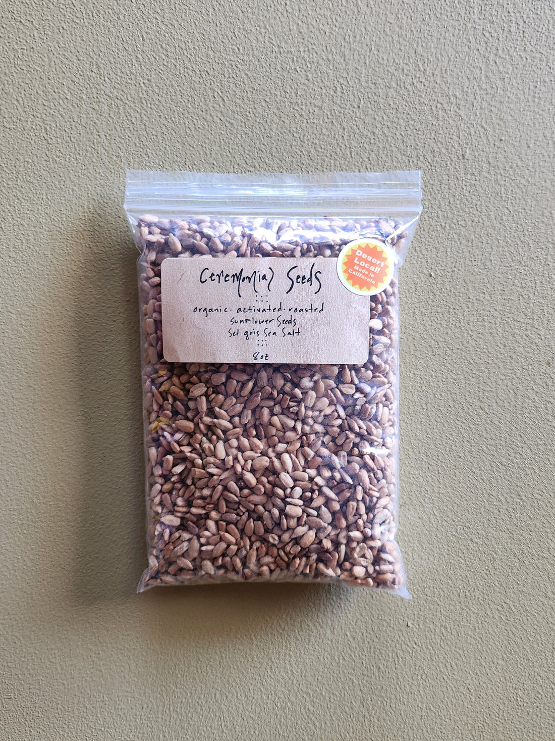 A clear plastic bag of Ceremonial Seeds organic roasted sunflower seeds. featuring a kraft paper label and a small orange circular sticker, set against a textured beige background.