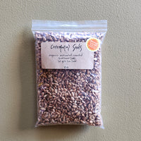 A clear plastic bag of Ceremonial Seeds organic roasted sunflower seeds. featuring a kraft paper label and a small orange circular sticker, set against a textured beige background.