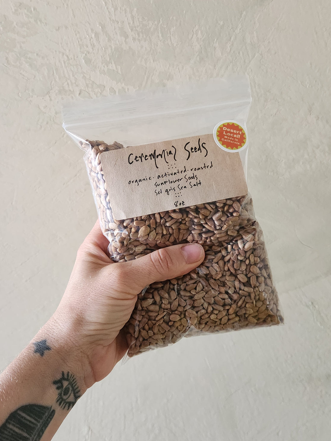 A hand holds a clear plastic bag filled with brown sunflower seeds against a textured off-white wall. The bag has a kraft paper label reading &