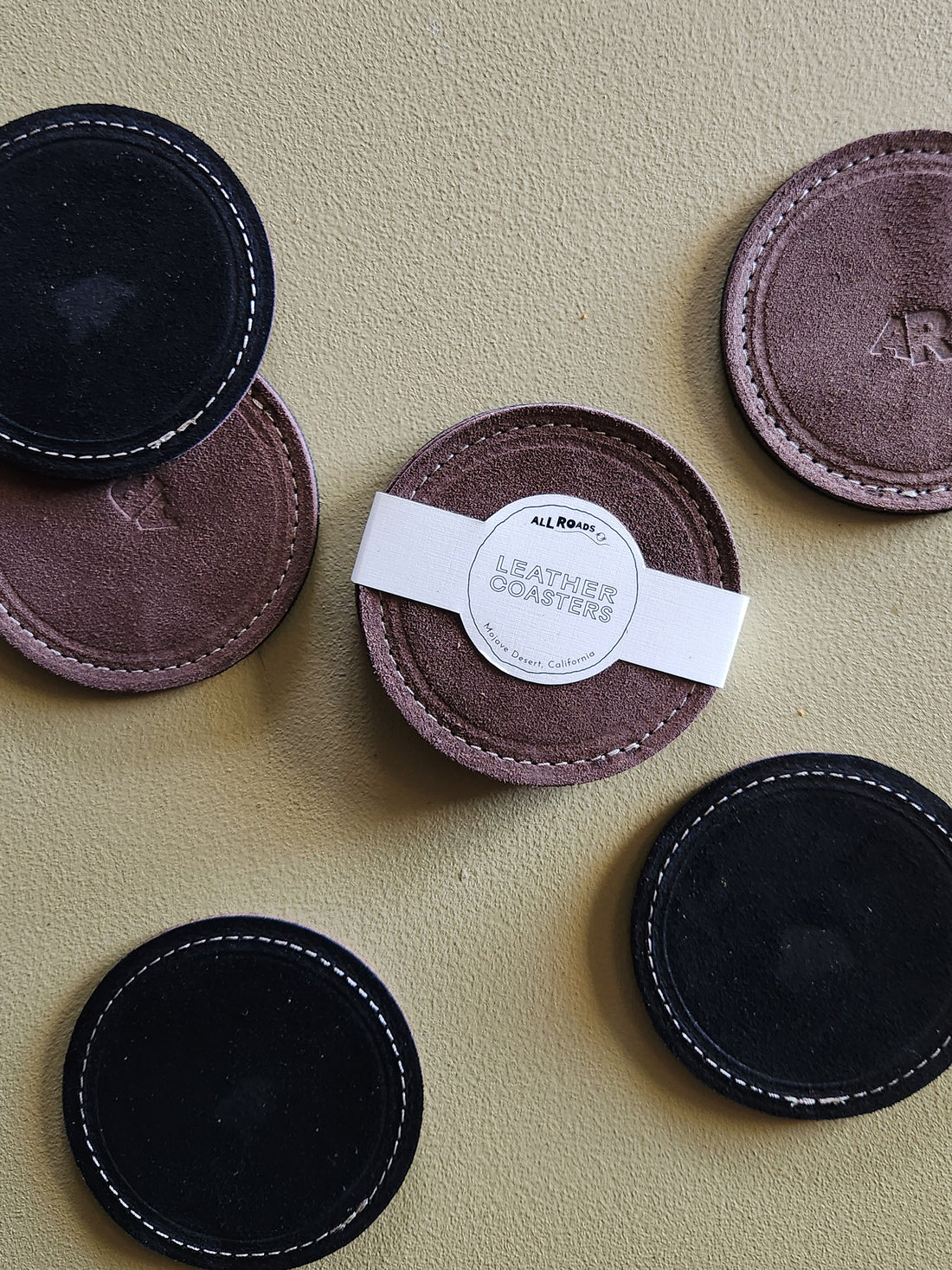 Six round leather coasters in black and muted mauve-brown tones arranged on a textured beige surface. One coaster has a white paper band labeled LEATHER COASTERS&