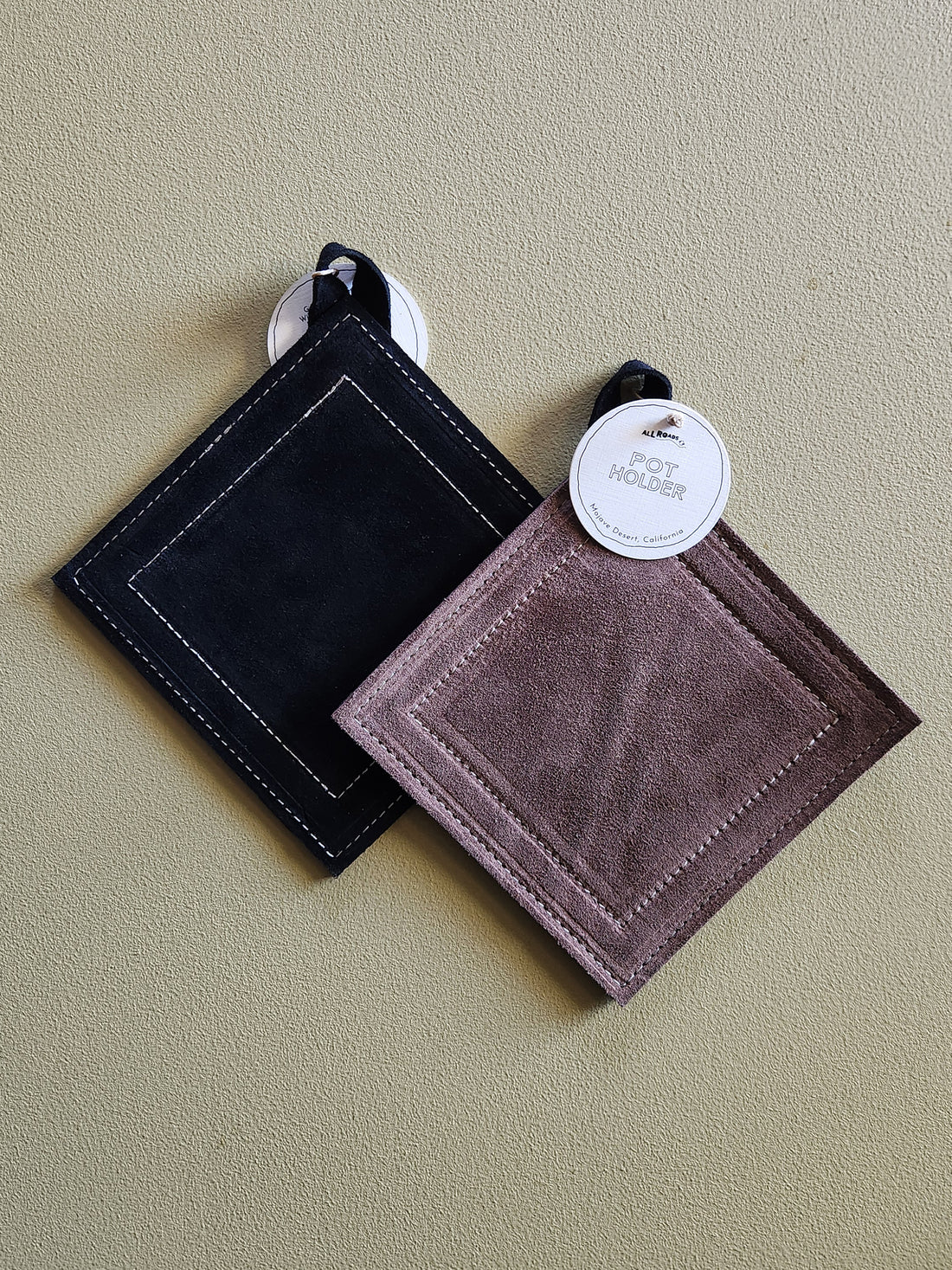 Two square suede-like potholders with stitched borders hang on a beige wall. One is black and the other is a muted mauve-brown, both with white hanging loops and round white product tags.