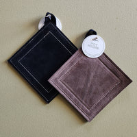 Two square suede-like potholders with stitched borders hang on a beige wall. One is black and the other is a muted mauve-brown, both with white hanging loops and round white product tags.